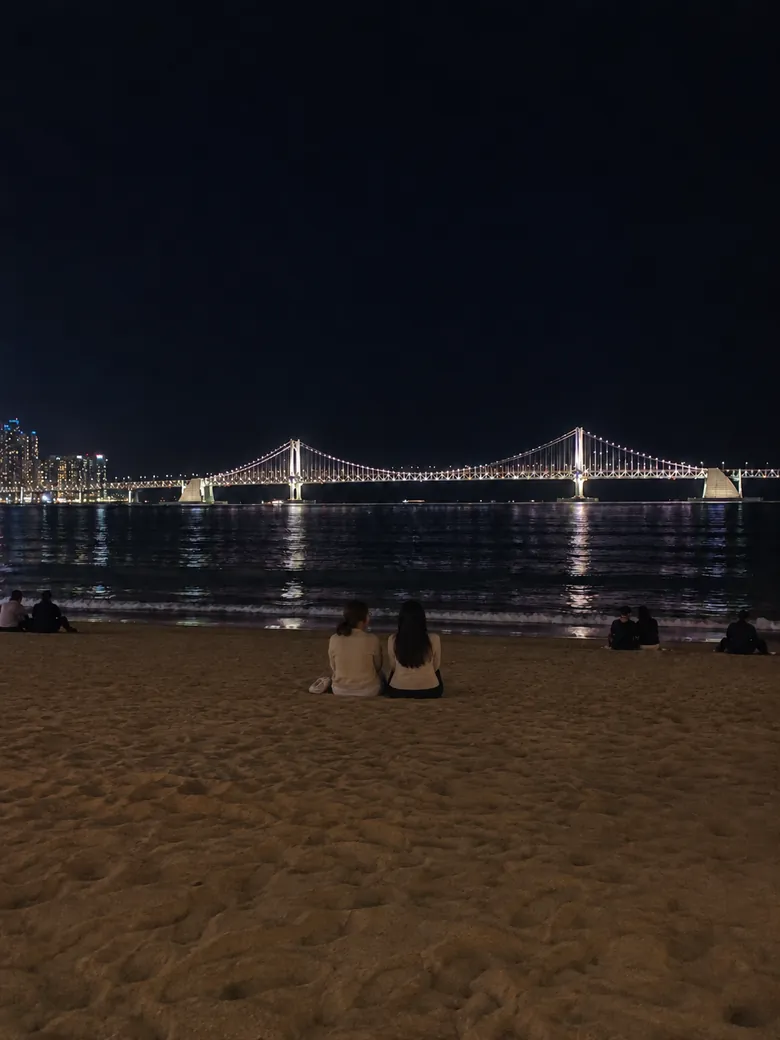 夜の広安里海水浴場に座り、ライトアップされた広安大橋を眺める人たち
